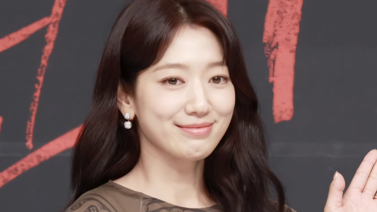 Park Shin-hye de cabelo longo escuro sorri e acena em coletiva de imprensa, diante de painel escuro com letras vermelhas