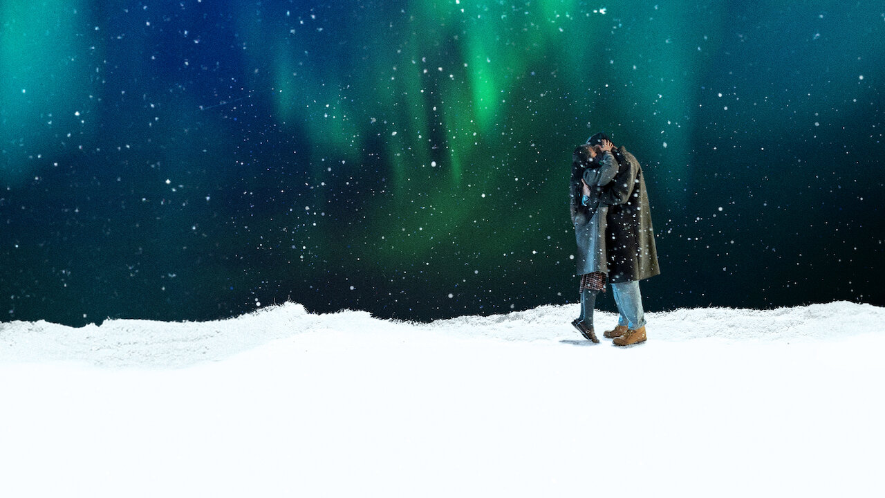 Casal abraçado de casacos sobre a neve, sob um céu noturno com aurora boreal verde e flocos de neve caindo
