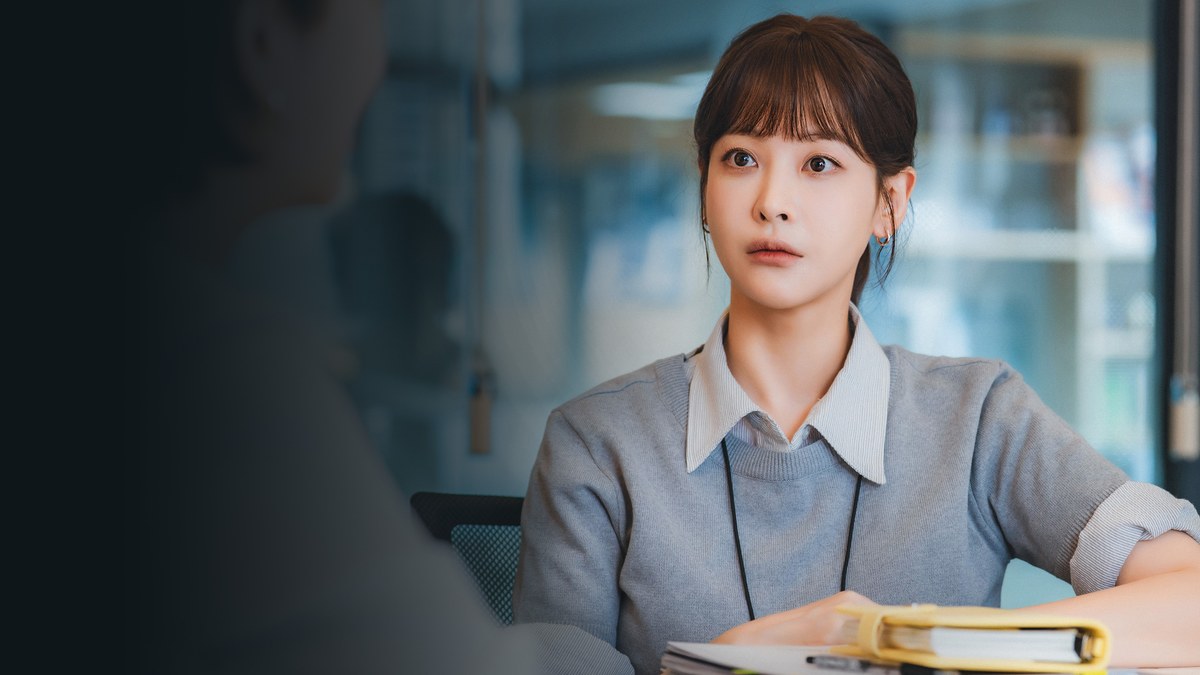 Arte de divulgação de Positively Yours na Viki: mulher de cabelo curto com franja, blusa cinza com crachá no pescoço, sentada a uma mesa com uma pasta amarela à frente e o lado esquerdo escurecido