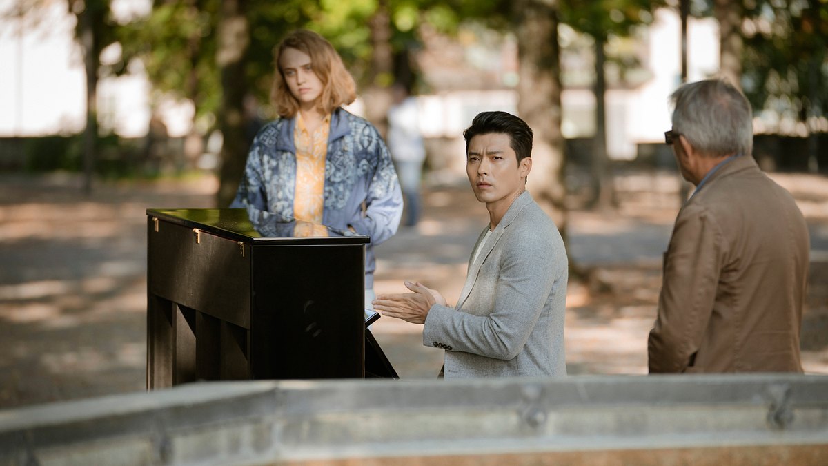 Homem de blazer cinza-claro sentado ao lado de um piano preto num parque arborizado, olhando para o lado, com uma jovem de jaqueta azul estampada atrás do piano e um senhor de casaco marrom à direita