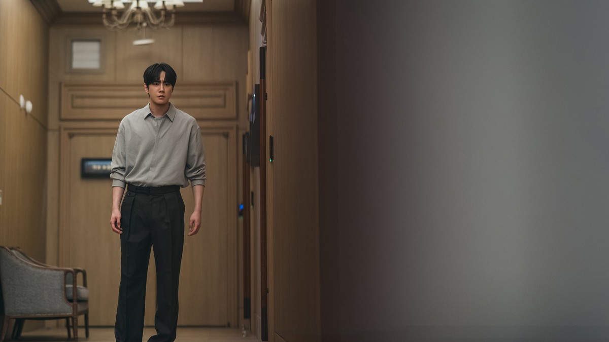Homem de cabelo escuro, camisa cinza e calça preta de pé num corredor com paredes de madeira clara e um lustre ao fundo, com a parte direita da imagem em sombra