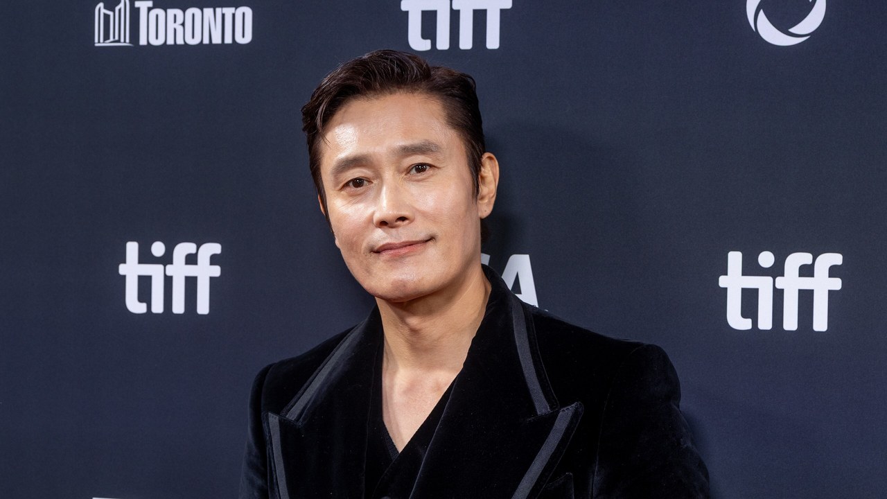 Lee Byung-hun, de cabelo castanho penteado para trás e blazer de veludo preto, sorri de leve diante do painel azul-escuro do Festival de Toronto