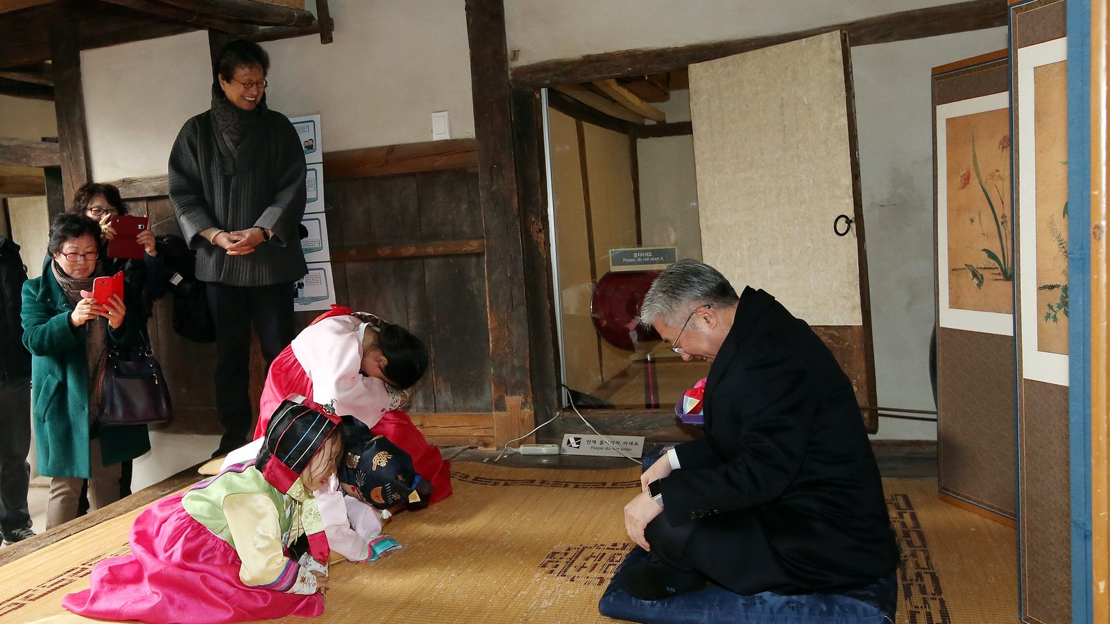 Duas crianças de hanbok curvadas no chão de esteira fazendo reverência diante de um homem de terno sentado numa almofada azul, numa casa tradicional