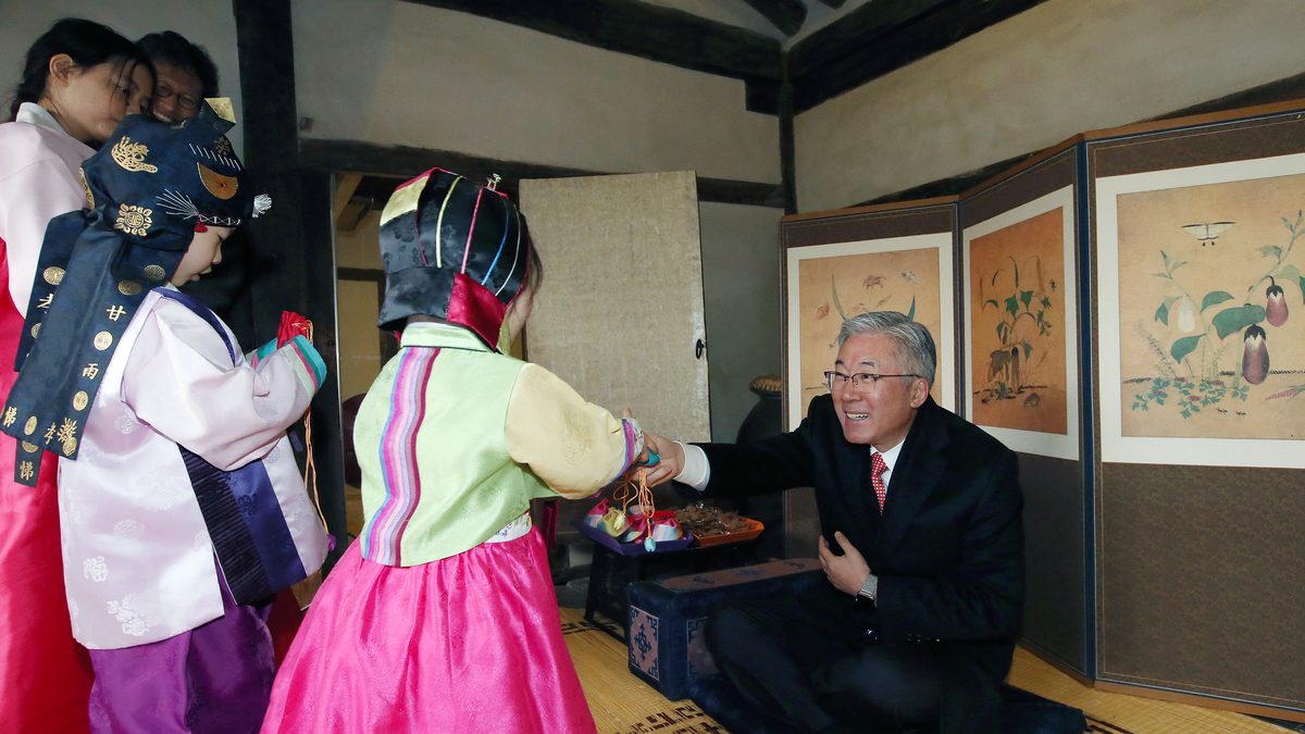 Menina de hanbok rosa e verde com chapéu tradicional estende a mão para um homem de terno agachado diante de um biombo pintado
