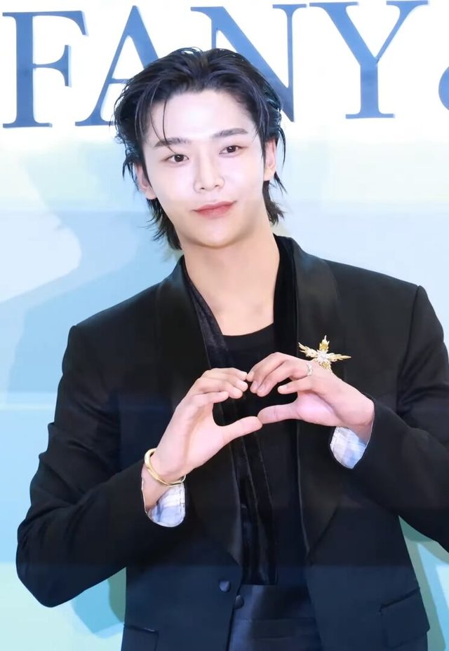 Rowoon de terno preto fazendo coração com as mãos diante de painel azul-claro