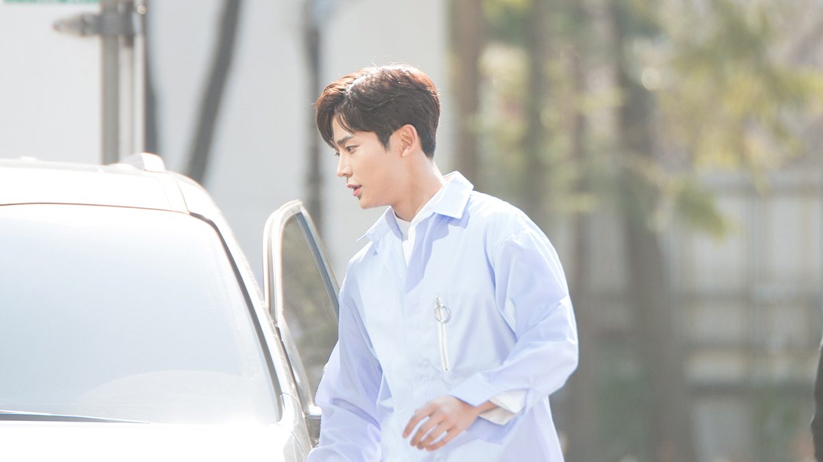 Rowoon de perfil, de camisa azul-clara, ao lado de um carro branco de porta aberta numa rua arborizada