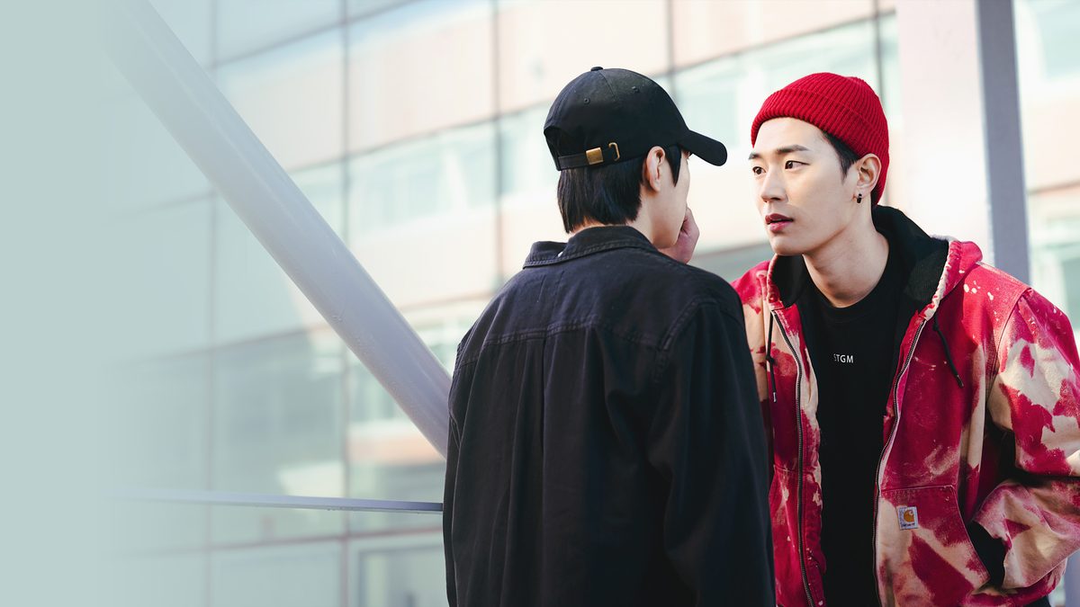 Dois rapazes frente a frente diante de uma fachada de vidro, um de costas com boné preto e camisa preta, o outro de gorro vermelho e jaqueta vermelha manchada, olhando para ele com expressão séria