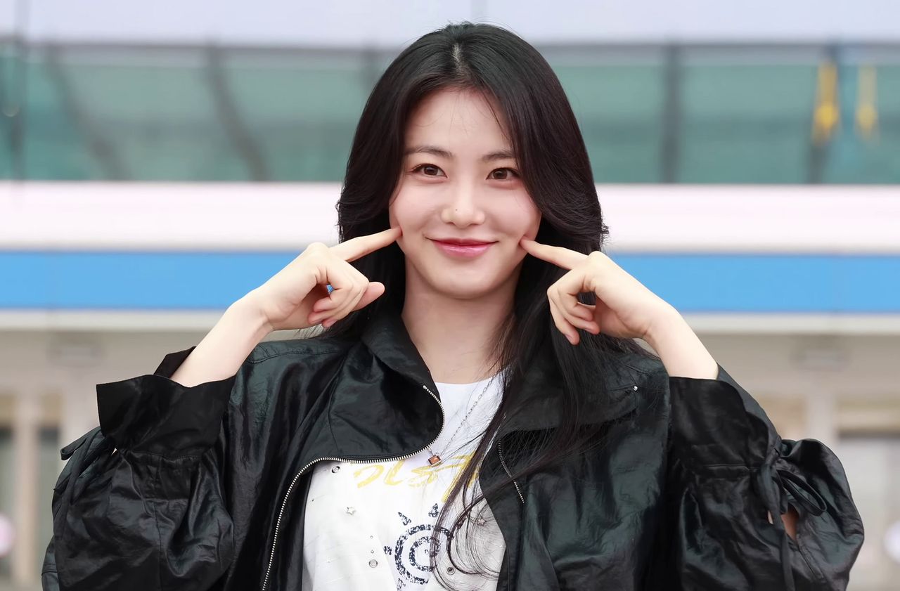 Mulher de cabelo preto longo, jaqueta preta bufante e camiseta branca sorrindo com os dedos indicadores nas bochechas, diante de uma fachada envidraçada