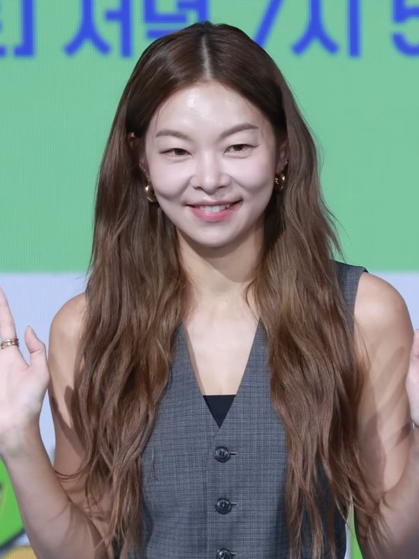 Song Hae-na de cabelo longo e colete cinza, sorrindo e acenando com as duas mãos
