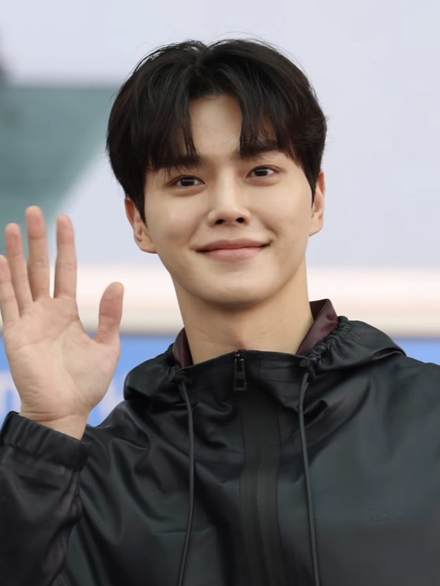Song Kang de jaqueta preta acenando e sorrindo em aeroporto