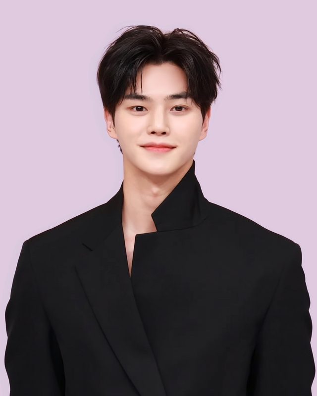 Song Kang sorrindo, de terno preto, diante de painel lilás numa coletiva de imprensa
