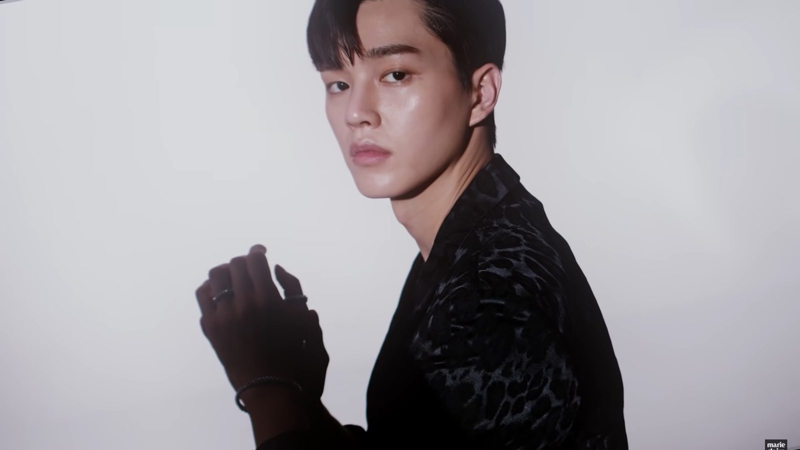 Song Kang olha por cima do ombro, de blazer preto estampado, com a mão de anéis erguida diante do peito, em fundo cinza de estúdio