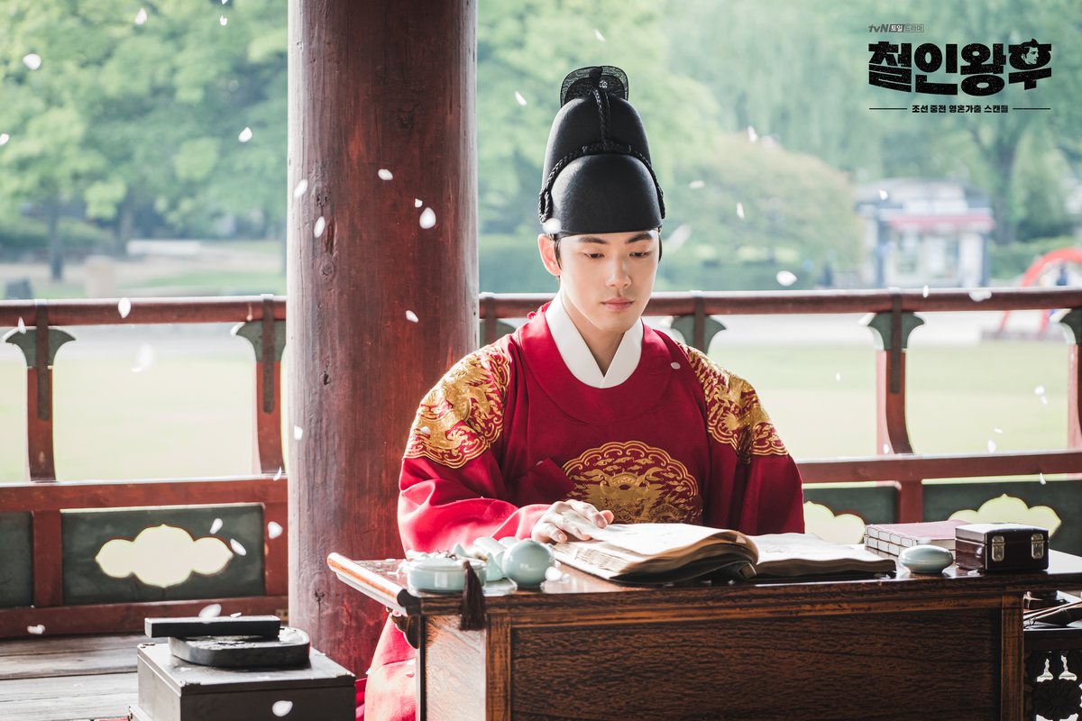 Kim Jung-hyun como Cheoljong, de manto vermelho com bordados dourados e chapéu preto, lê um livro numa mesa baixa dentro de um pavilhão aberto para o jardim