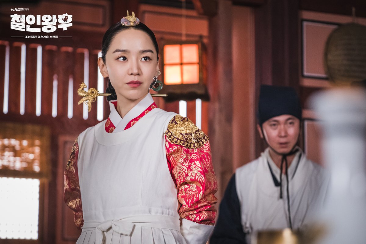 Shin Hye-sun como So-yong, de avental branco sobre o traje vermelho e dourado, com um enfeite de fênix no cabelo; ao fundo, um homem de roupa branca e chapéu escuro