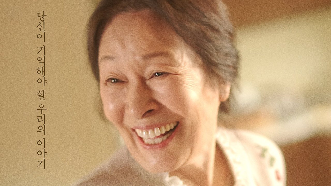 Close de Kim Hye Ja, senhora de cabelo grisalho preso, sorrindo de boca aberta, de blusa branca com bordado de flores, com a frase de chamada do pôster em coreano na lateral esquerda
