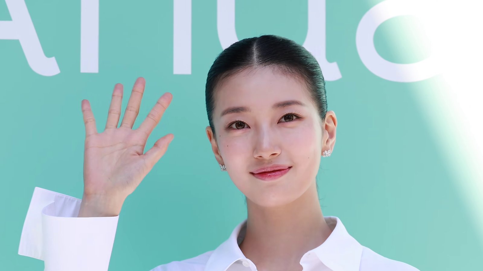 Suzy de camisa branca e cabelo preso sorri e acena diante de um painel verde-água
