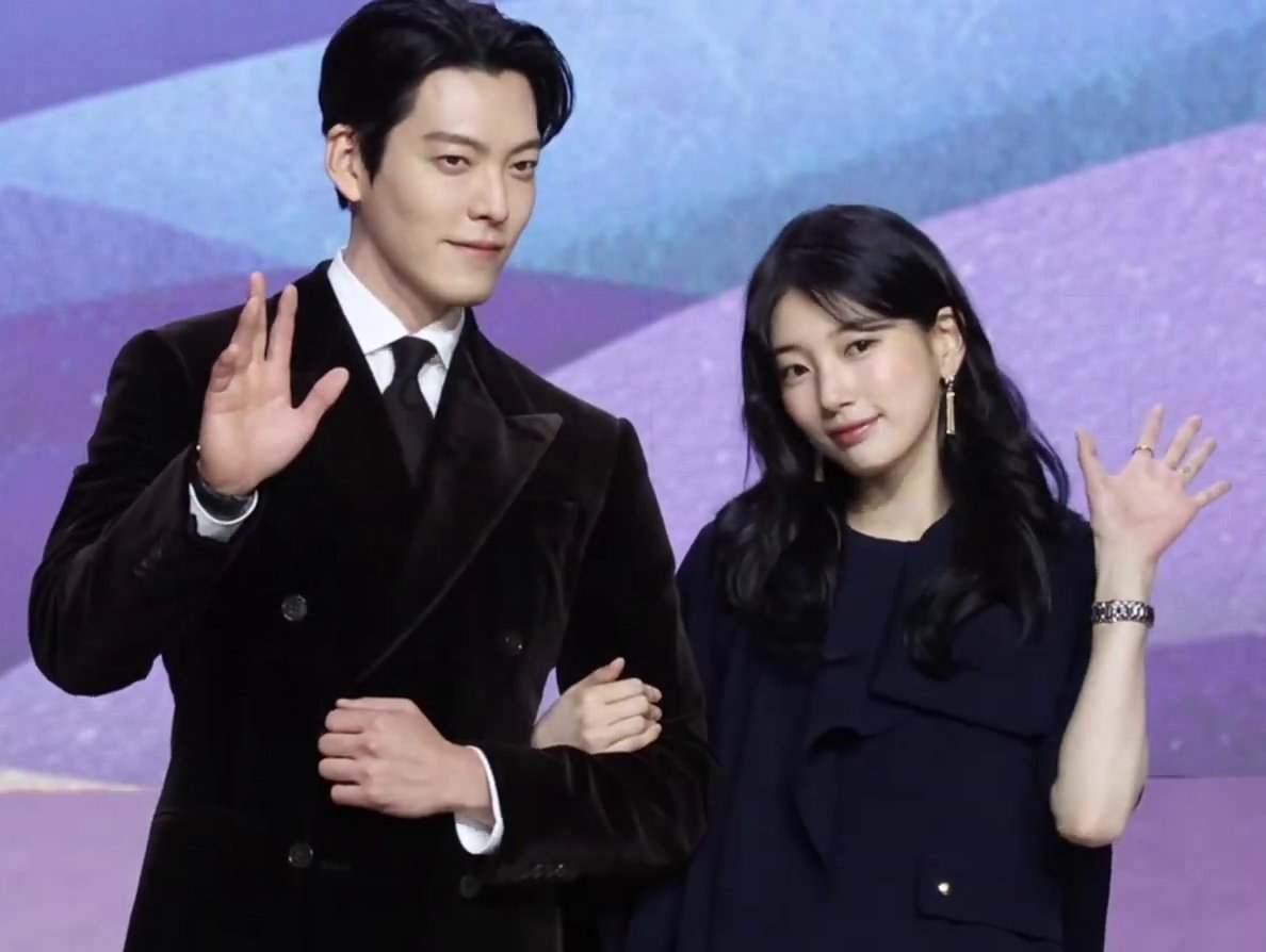 Kim Woo-bin de terno escuro e Suzy de vestido azul-marinho acenam de braços dados diante de painel lilás