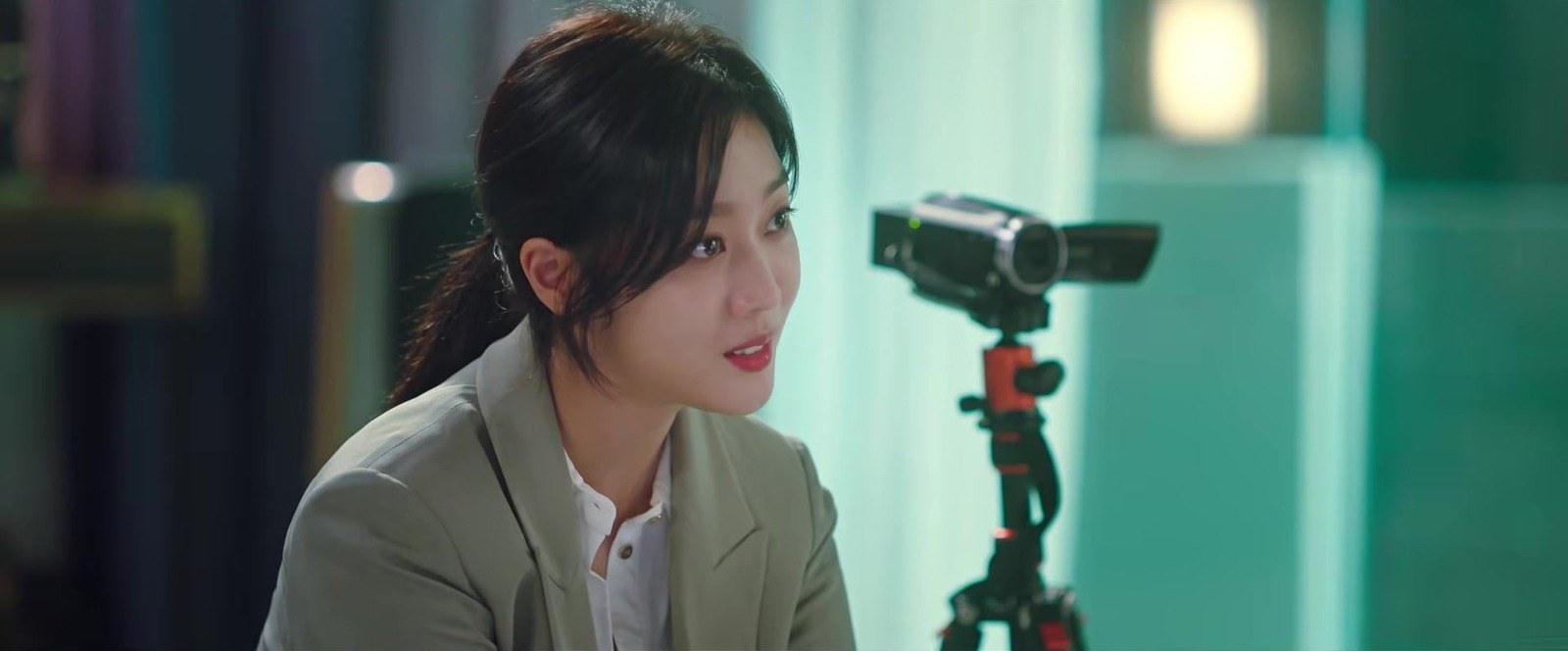 Mulher de cabelo preso e blazer verde-claro conversa diante de uma câmera de vídeo num tripé vermelho, em quadro do trailer oficial de Tale of the Nine Tailed
