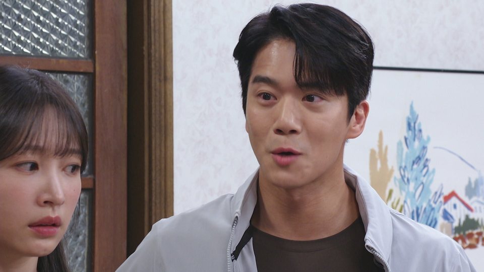 Homem de jaqueta branca sobre camiseta marrom fala com expressão animada, ao lado de uma mulher de cabelo escuro em primeiro plano, numa sala com um quadro colorido na parede