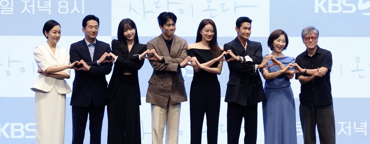 Oito atores de Tempero do Amor fazem coração com as mãos, lado a lado, diante do painel da KBS com o nome da novela