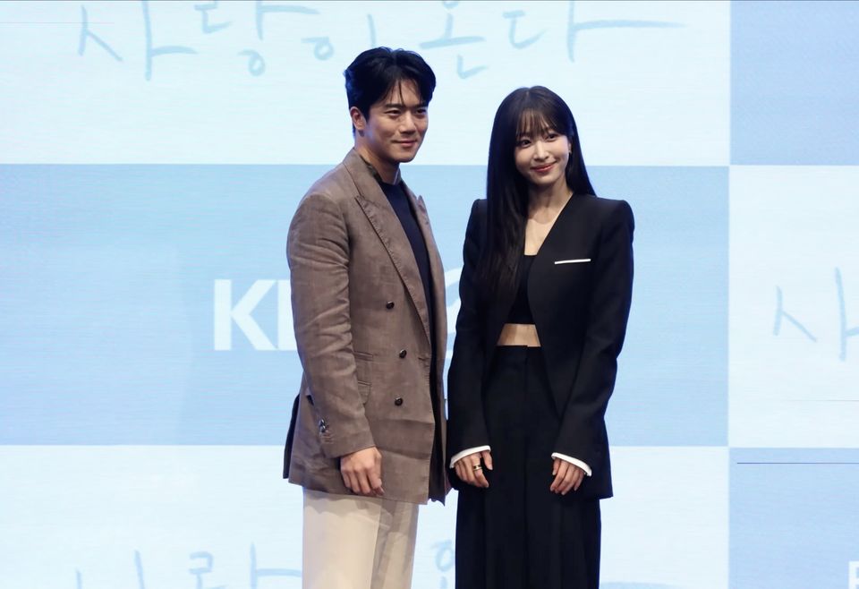 Ha Seok-jin, de paletó xadrez marrom, e Hani, de terno preto, posam juntos diante do painel azul da coletiva