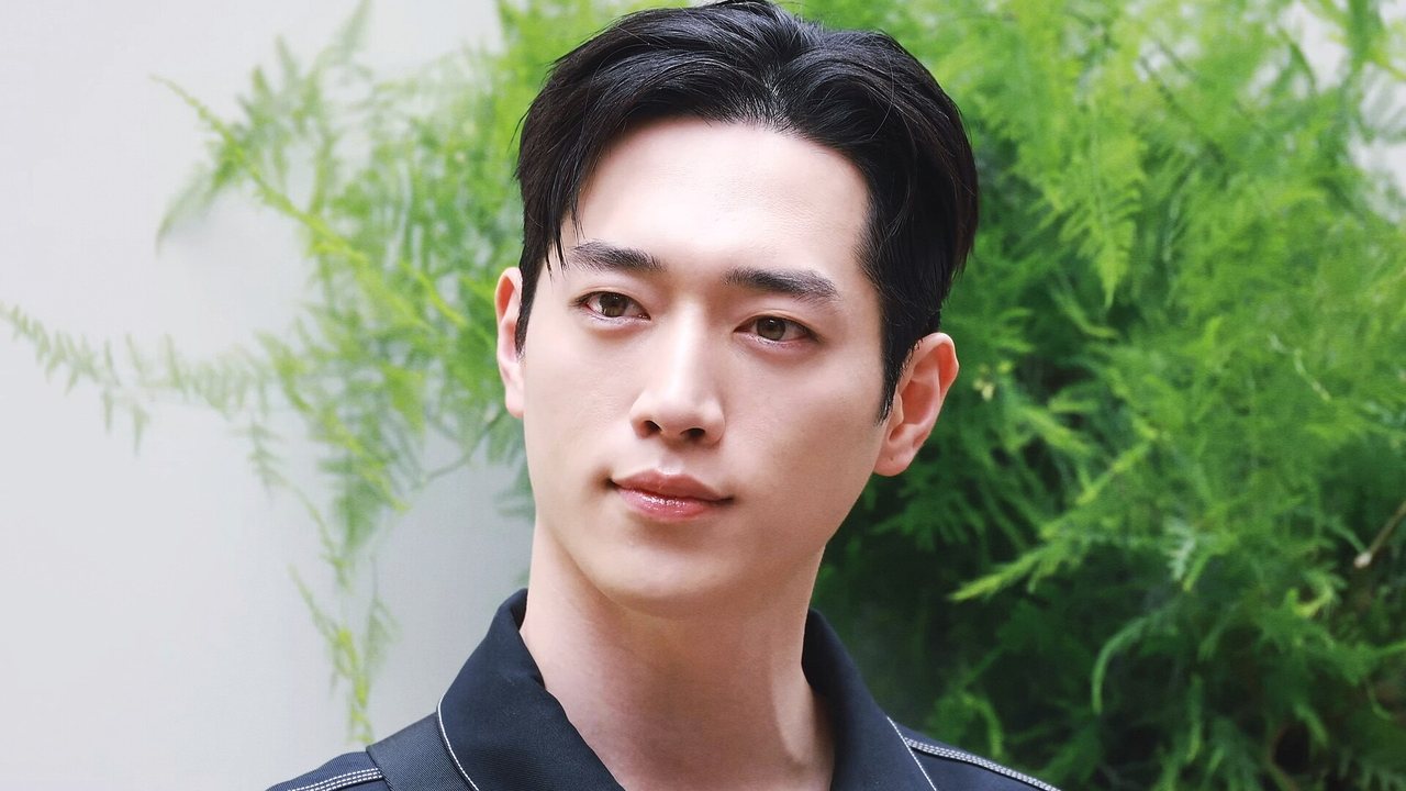 Seo Kang-joon, de camisa escura, olha para a câmera diante de uma planta verde em evento