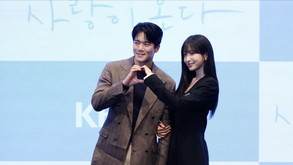 Ha Seok-jin, de blazer marrom, e Hani, de blazer preto, fazem um coração com as mãos diante do painel azul da KBS na coletiva de Tempero do Amor
