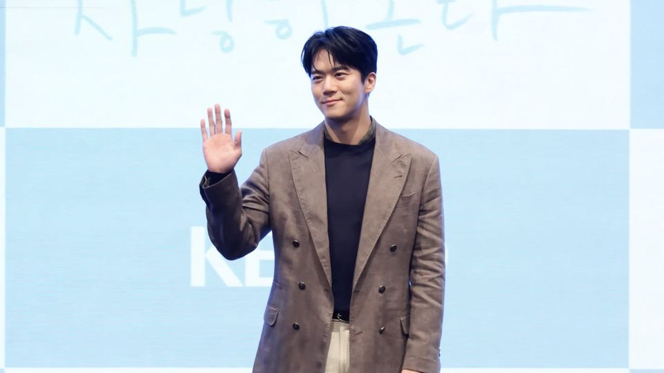 Ha Seok-jin, de blazer marrom e camisa preta, acena com a mão direita diante do painel da KBS na coletiva de Tempero do Amor