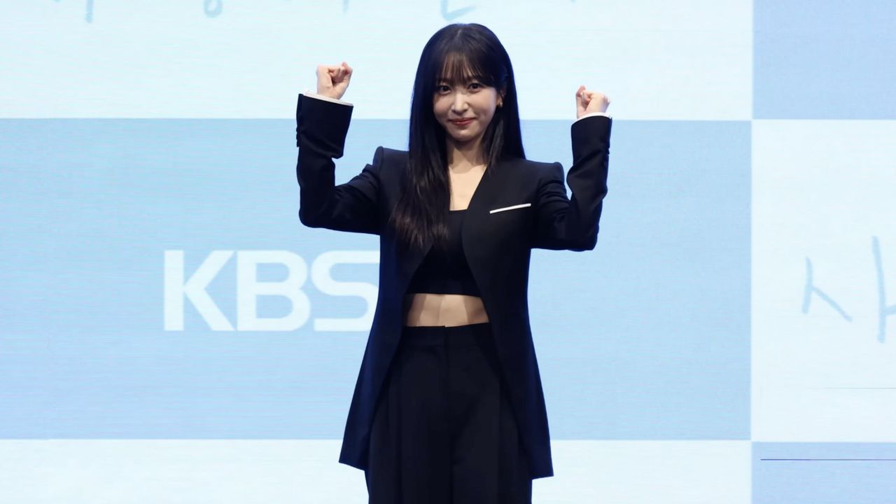 Hani, de blazer e calça pretos, sorri com os dois punhos erguidos diante do painel azul da KBS na coletiva de Tempero do Amor