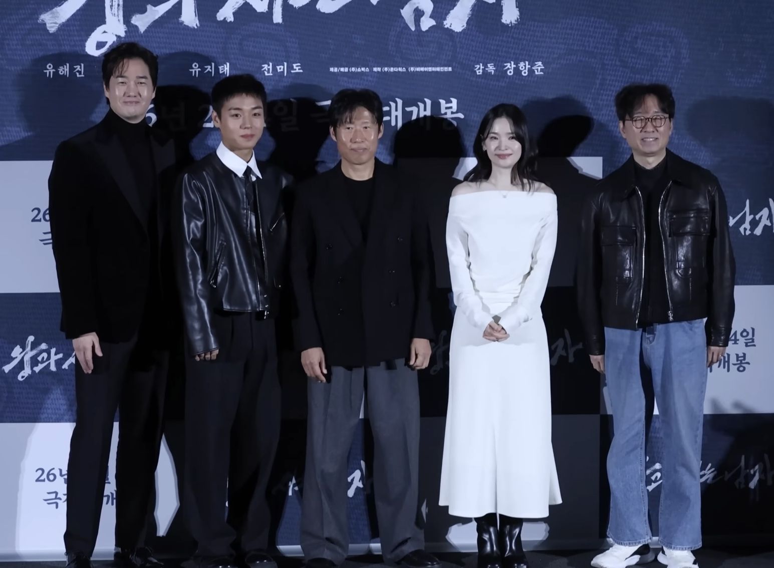 Cinco pessoas lado a lado diante de um painel azul escuro: um homem de roupa preta e gola alta, um jovem de jaqueta de couro, Yoo Hae Jin ao centro de blazer preto, uma mulher de vestido branco e um homem de óculos e jaqueta de couro
