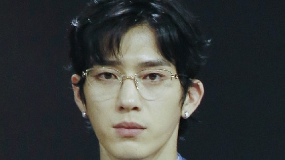 Jing Boran de óculos de armação dourada e cabelo escuro, sério, diante de fundo preto