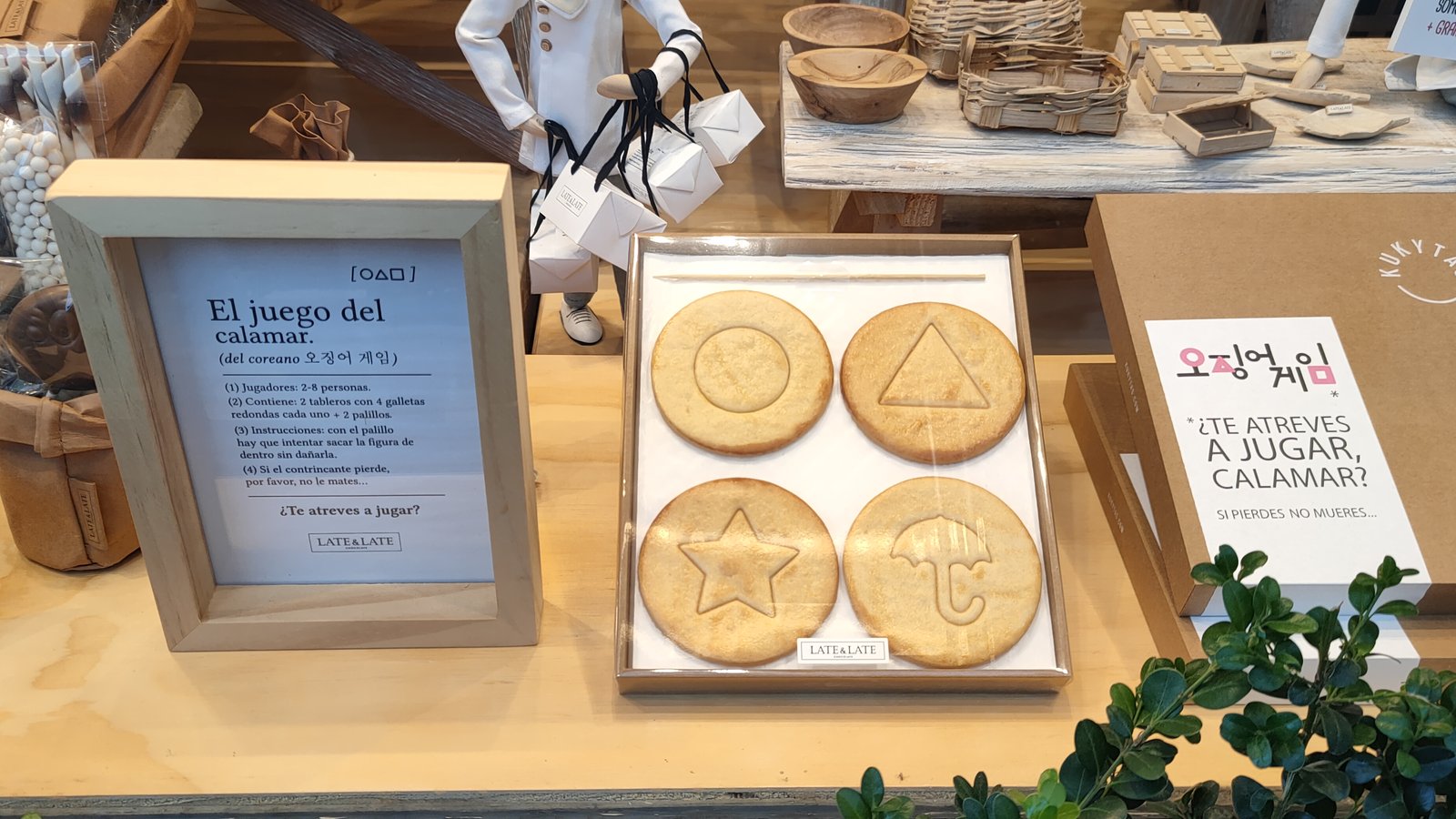 Vitrine na Espanha com um quadro impresso com o título El juego del calamar e o nome em coreano, quatro biscoitos com formas de círculo, triângulo, estrela e guarda-chuva e uma caixa com 오징어 게임