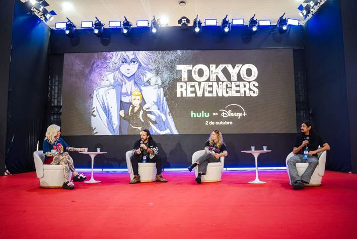 Palco com quatro pessoas sentadas em poltronas brancas diante de um telão com a arte de Tokyo Revengers, as marcas hulu e Disney+ e a data 2 de outubro, com plateia em primeiro plano