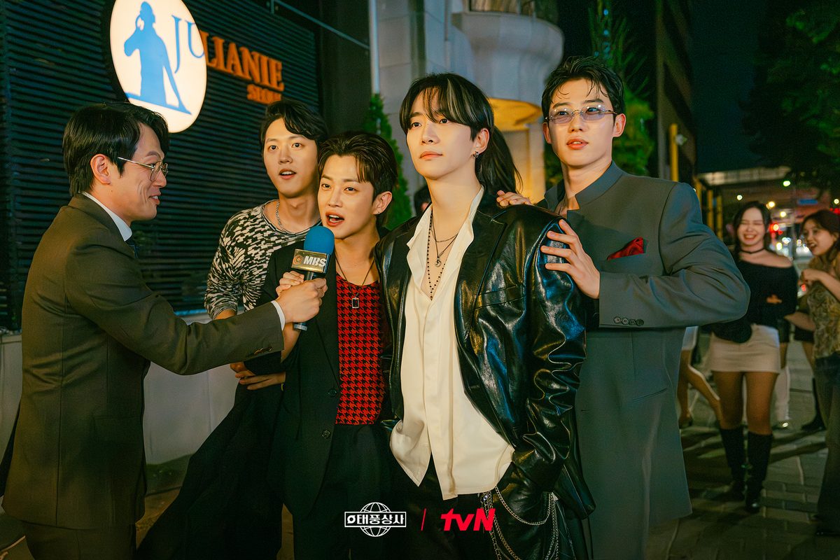 À noite, numa rua com letreiros, um repórter estende um microfone a um jovem enquanto Lee Jun-ho, de jaqueta de couro preta e camisa branca, posa com amigos de roupas coloridas