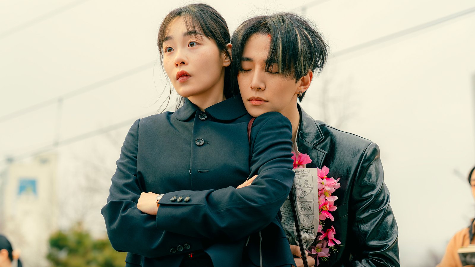 Lee Jun-ho, de jaqueta de couro e com um buquê de flores cor-de-rosa, apoia o queixo no ombro de Kim Min-ha, de casaco escuro e braços cruzados, numa calçada