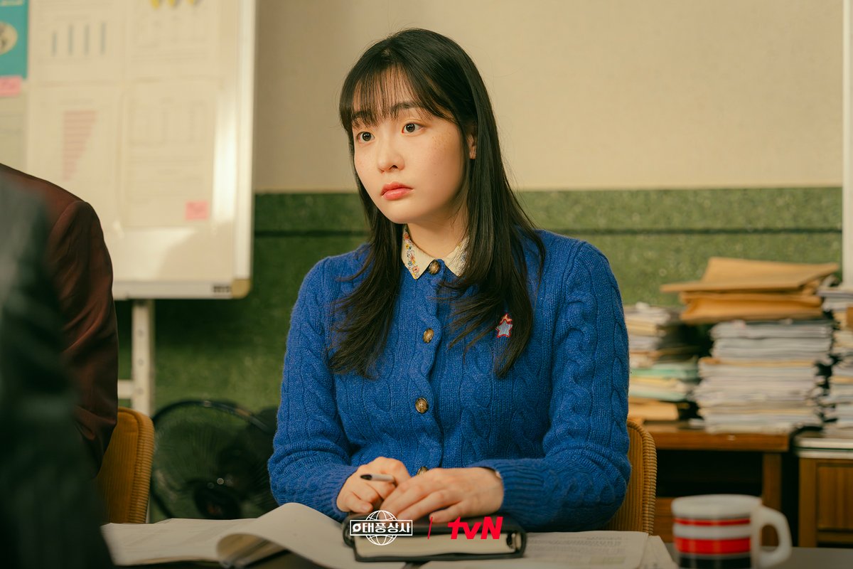 Kim Min-ha como Oh Mi-seon, de cardigã azul e gola bordada, sentada a uma mesa de escritório com papéis empilhados ao fundo