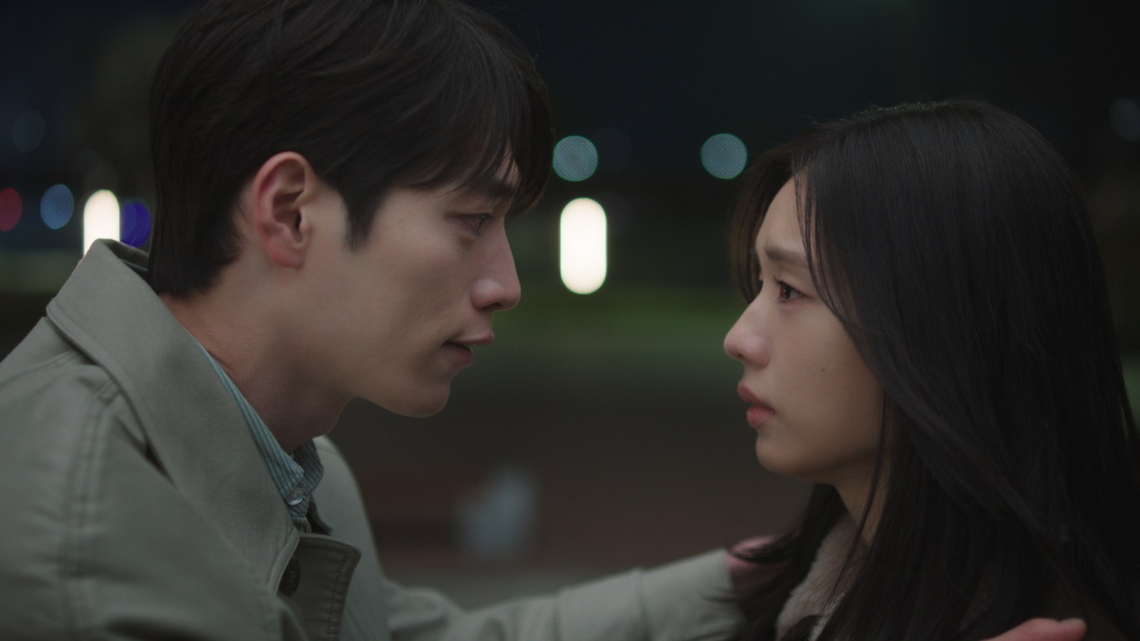 Seo Kang-joon de casaco bege e Ahn Eun-jin de cabelo solto, frente a frente à noite, com luzes desfocadas ao fundo
