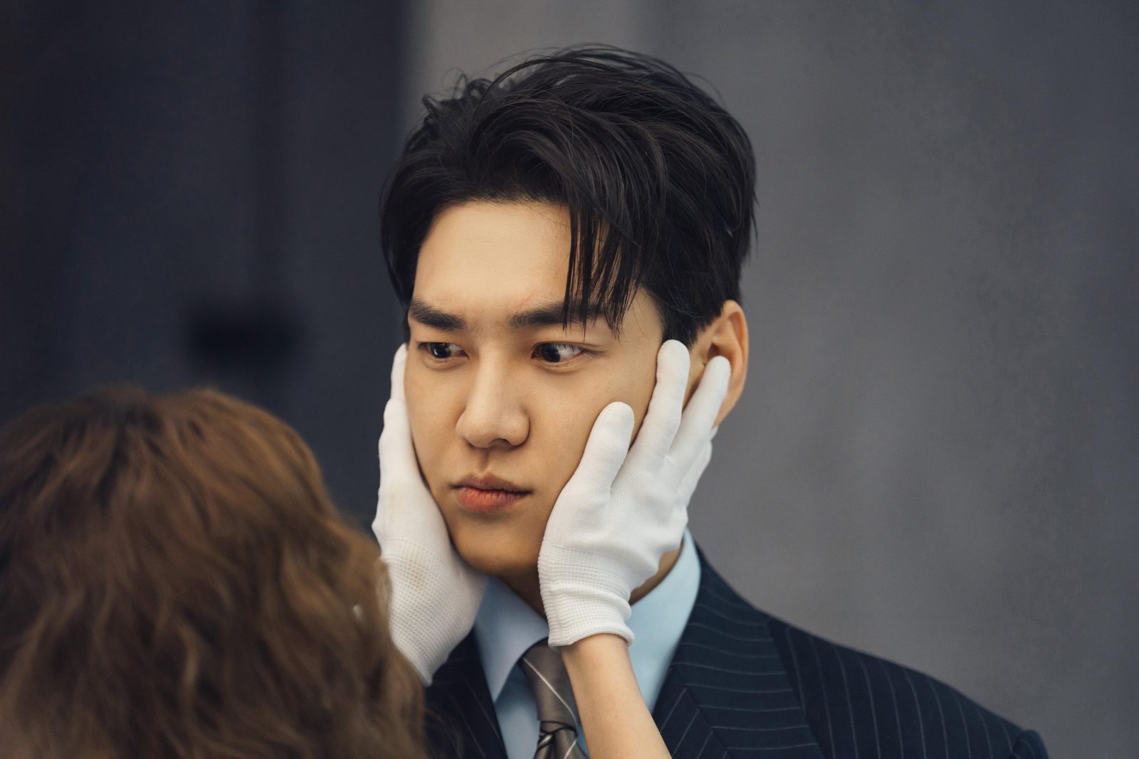 Kim Young Kwang de terno risca de giz tem o rosto segurado por mãos de luva branca, enquanto Chae Soo Bin aparece de costas em primeiro plano, em cena de Um Choque de Amor