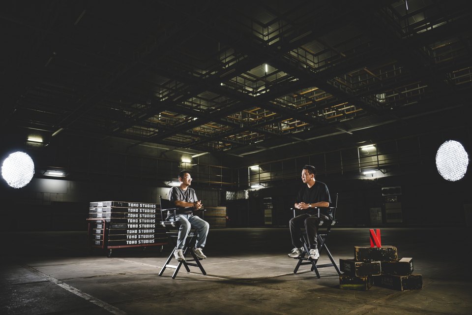 Dois homens sentados em cadeiras de diretor num estúdio escuro e amplo, com refletores acesos, caixas com o nome Toho Studios e um logotipo da Netflix