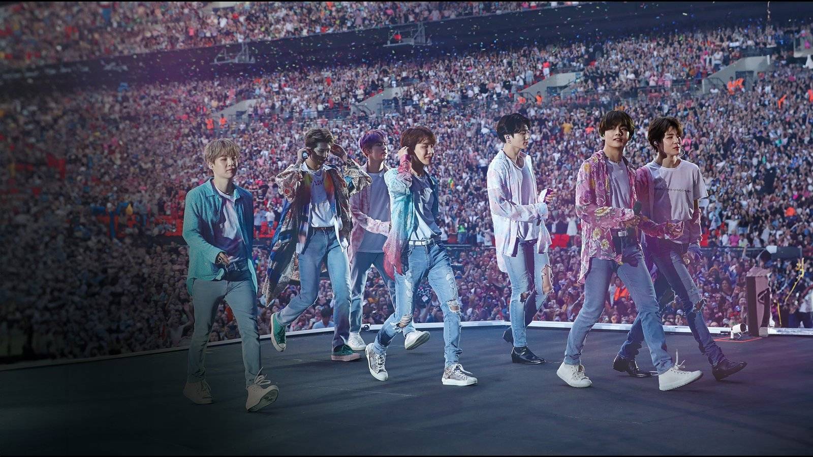 Os sete integrantes do BTS caminham lado a lado num palco diante de um estádio lotado, com confete no ar e luz roxa e azul