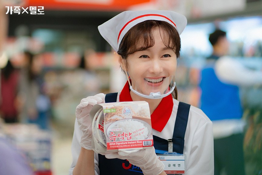 Kim Ji-soo como Geum Ae-yeon, de touca, avental e protetor facial transparente, sorri mostrando uma embalagem de arroz instantâneo num corredor de supermercado