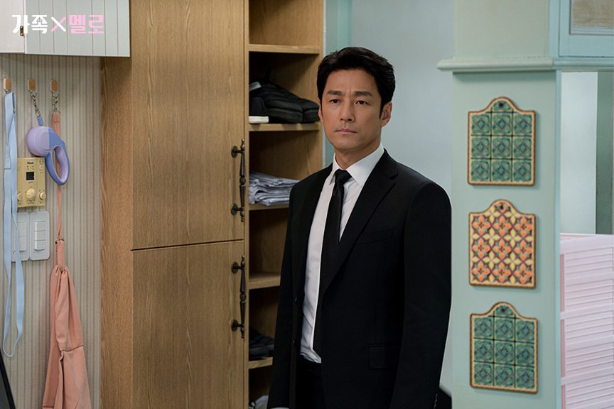 Ji Jin-hee como Byeon Moo-jin, de terno e gravata pretos, parado na entrada de um apartamento com armário de madeira e azulejos decorativos