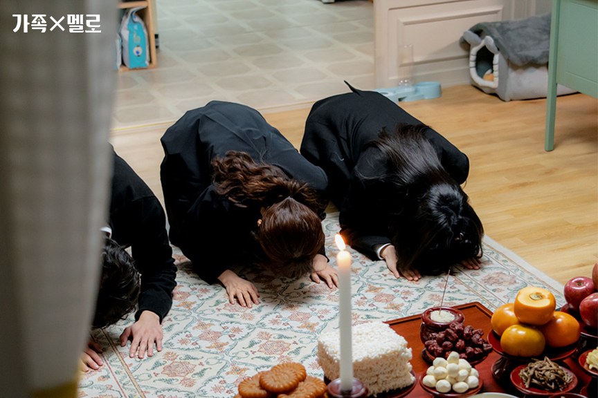 Família vestida de preto curvada no chão diante de uma mesa de ritual com vela acesa, frutas, bolinhos e bolo de arroz branco