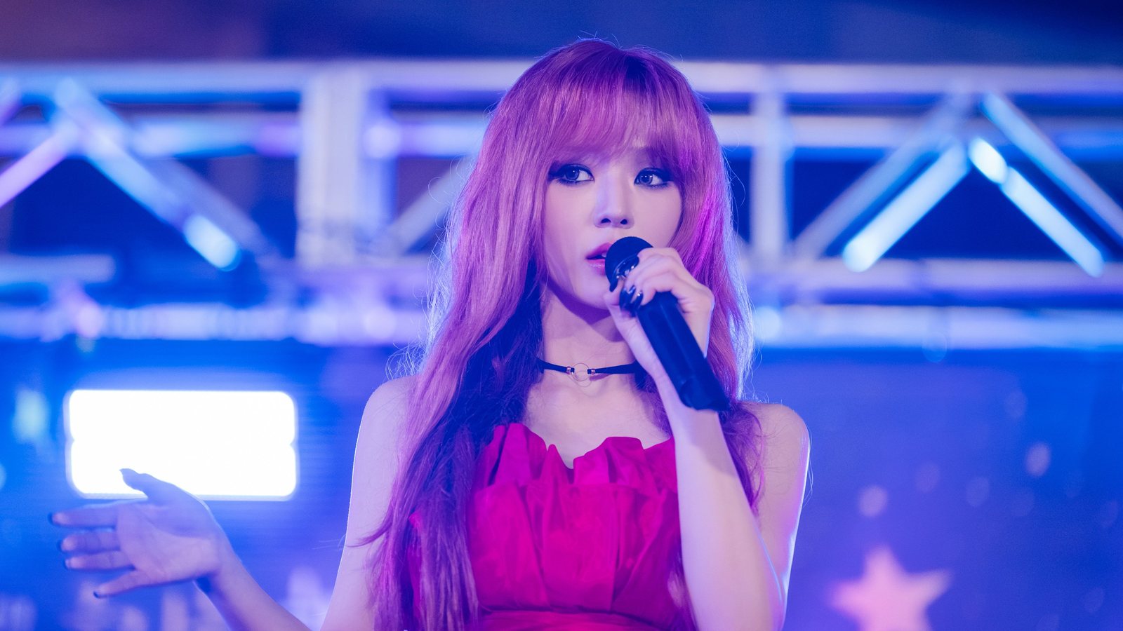Oh Se Young, de cabelo longo rosa-lilás e vestido pink, canta de microfone na mão direita diante de uma treliça de palco desfocada em azul