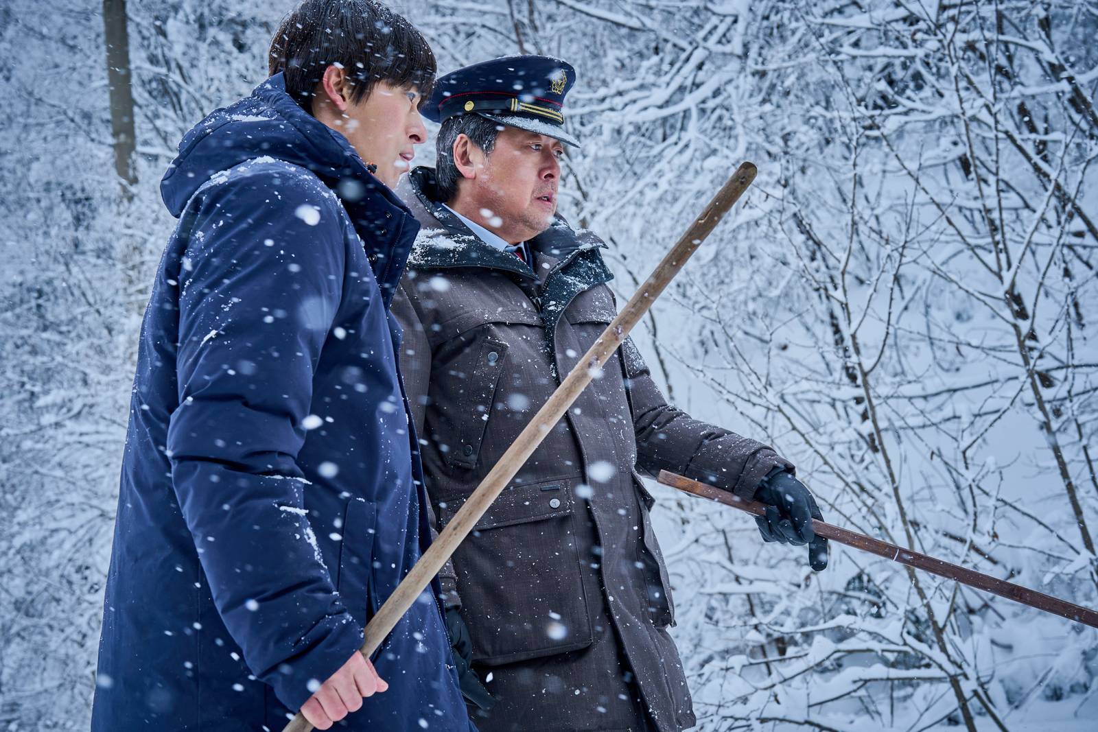 Dois homens caminham na neve em meio a árvores, um de parca azul-marinho e outro de quepe de ferroviário e casaco marrom, em cena de White Blast