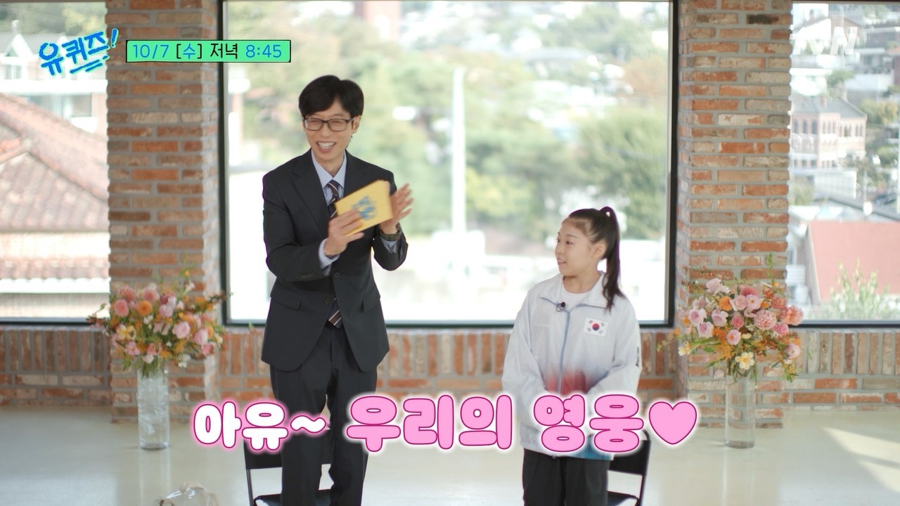 Yoo Jae-suk, de terno, segura um envelope amarelo ao lado da ginasta Hwang Seo-hyun, de agasalho branco da delegação coreana, numa sala de tijolos aparentes