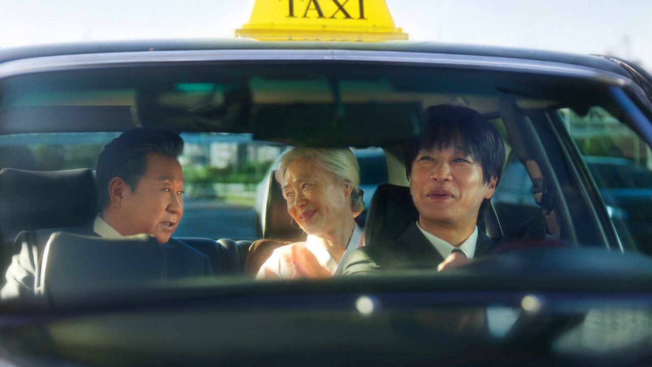 Homem de cabelo escuro e terno ao volante de um táxi preto sorri; no banco de trás, uma mulher idosa de cabelo branco e roupa rosa clara e um homem de terno conversam, vistos pelo para-brisa, com a placa amarela TAXI no teto